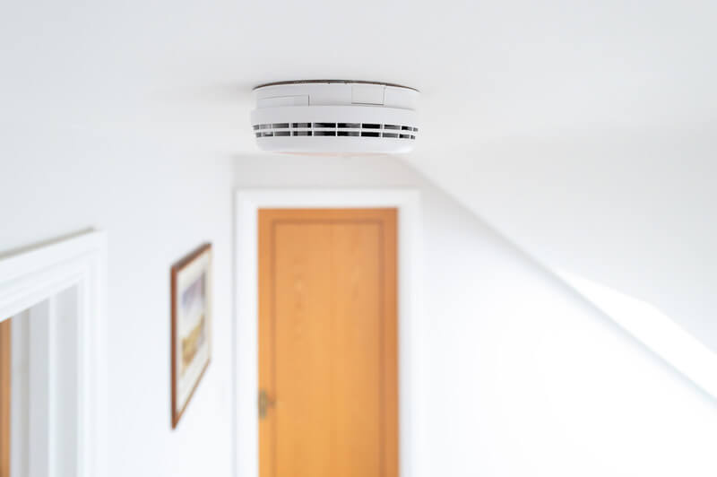 A smoke detector on the ceiling in a hallway in front of a door